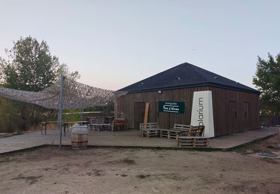 Bâtiment bas à bardage de bois vertical sous un toit sombre en croupe, portant une enseigne verte « Guinguette Les 2 Rives ». Devant, une terrasse en planches meublée de banquettes en palettes et d'un tonneau, sous une voile d'ombrage tendue vers les arbres.